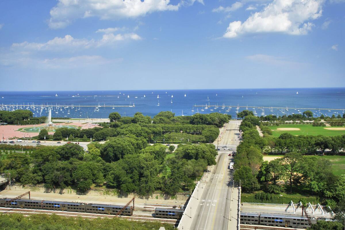 Un paisaje urbano costero con una vía de tren, áreas verdes y un puerto deportivo con barcos.