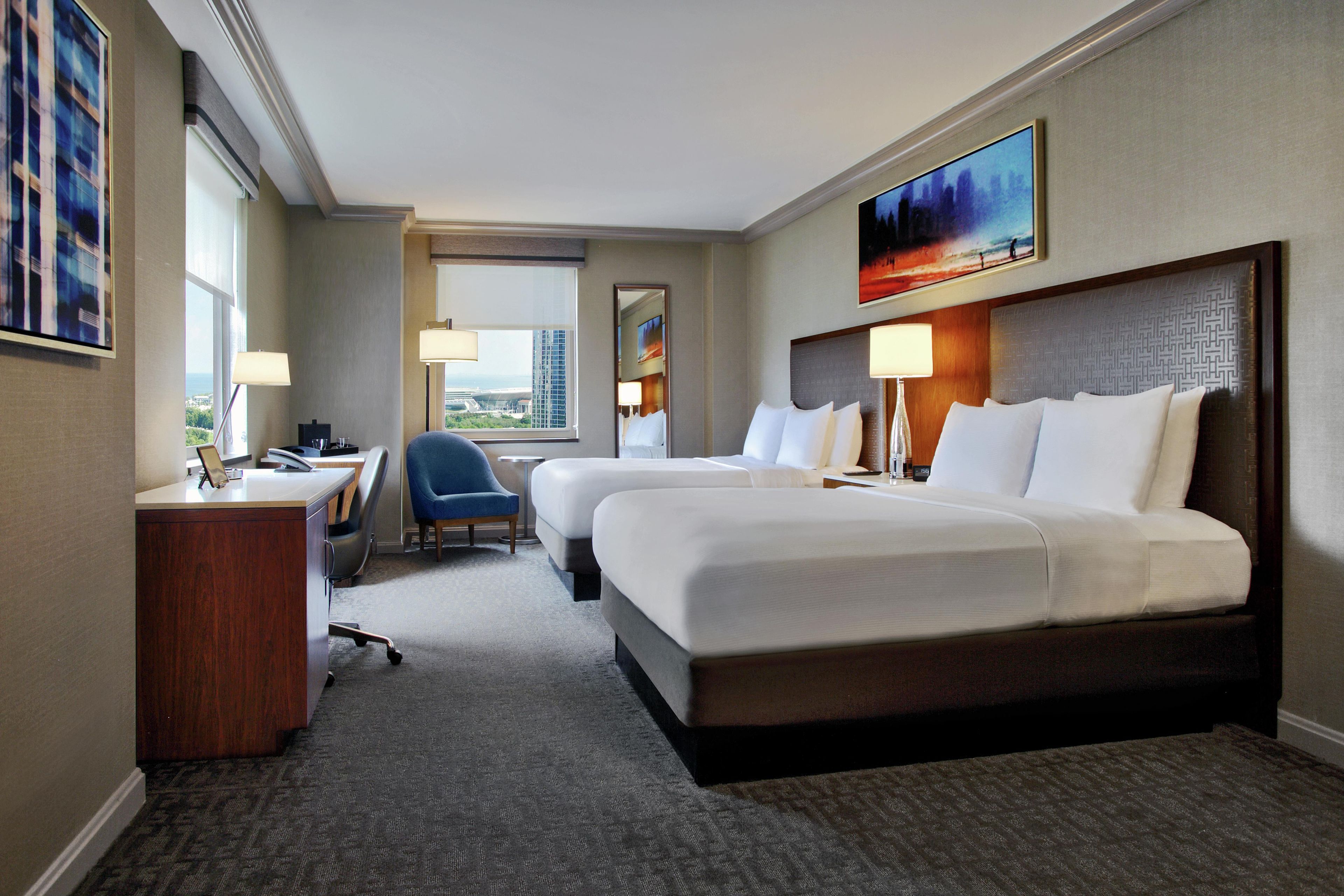 A hotel room with two beds, a desk, a chair, and a view of the cityscape.
