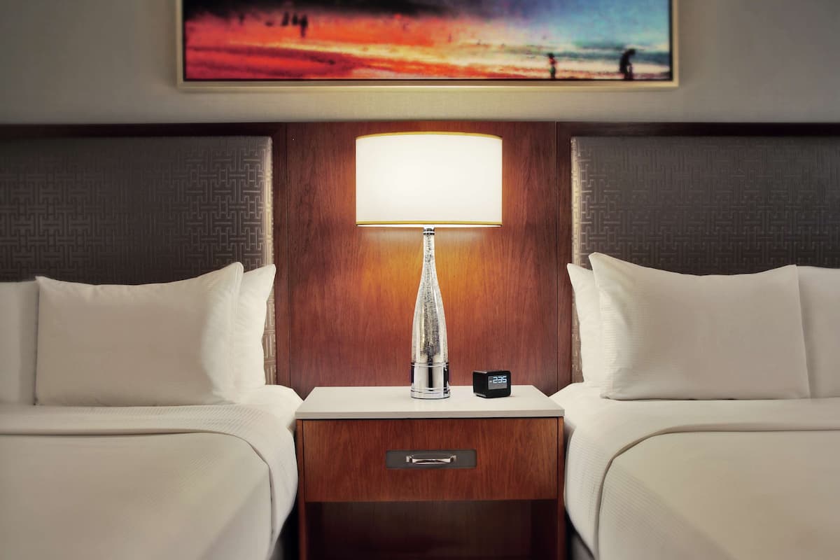 Una habitación de hotel con dos camas, una mesita de noche con lámpara, un reloj y un cuadro en la pared.