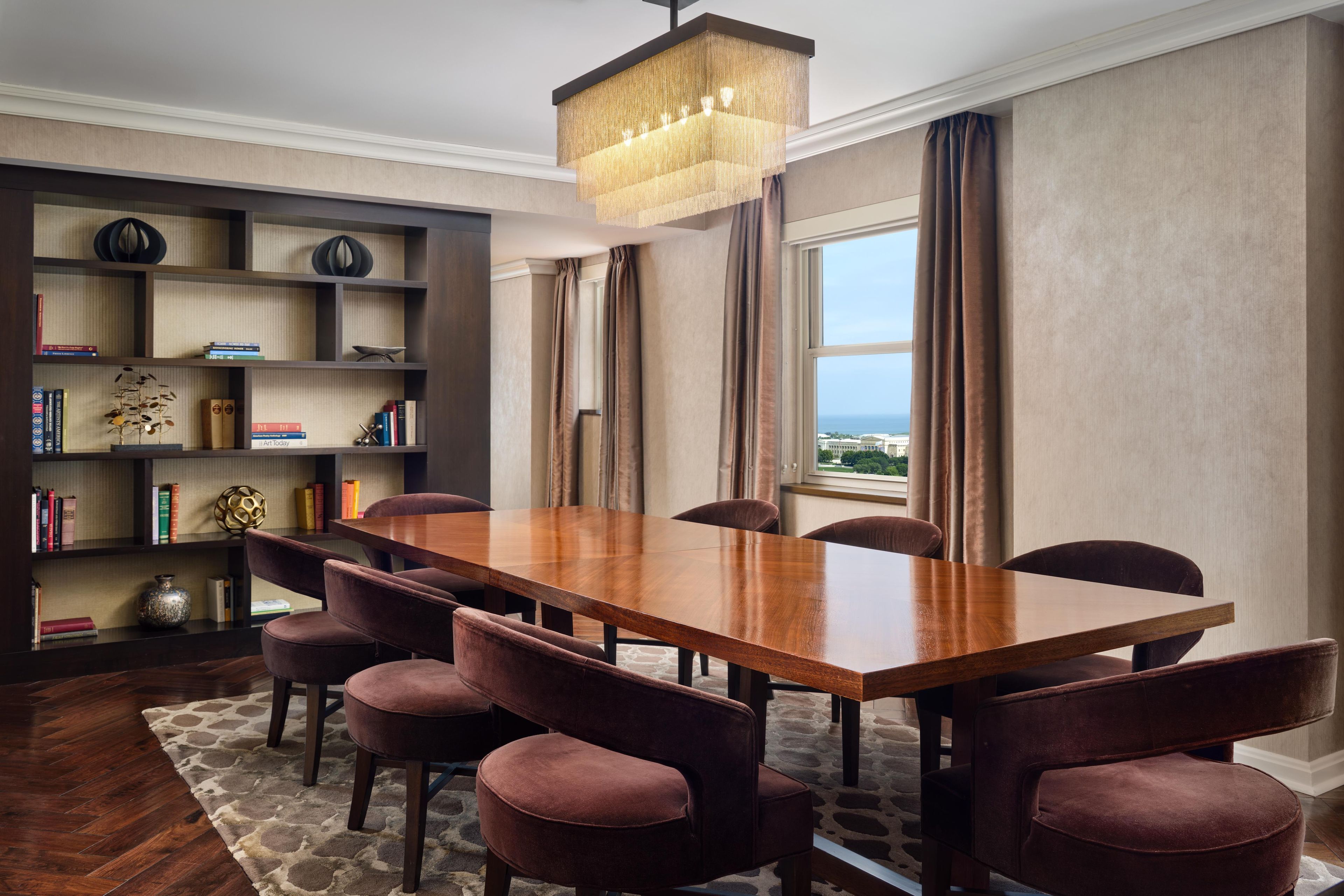 A dining area with a wooden table and six chairs, a bookshelf with various items, and a view through a window.