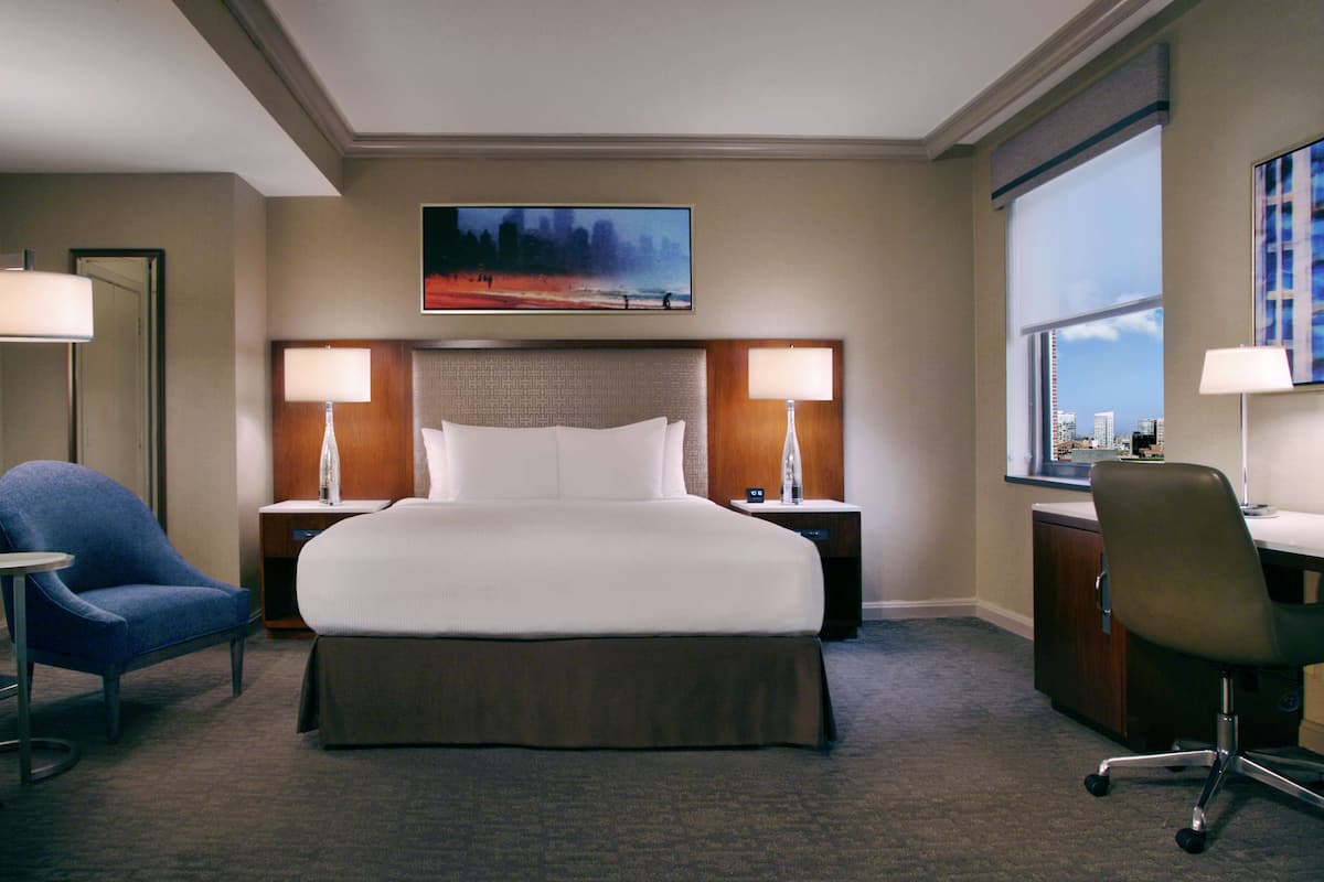 Una habitación de hotel con una cama grande, dos mesitas de noche con lámparas, un escritorio con una silla, una ventana con vistas a la ciudad y un cuadro sobre la cama.