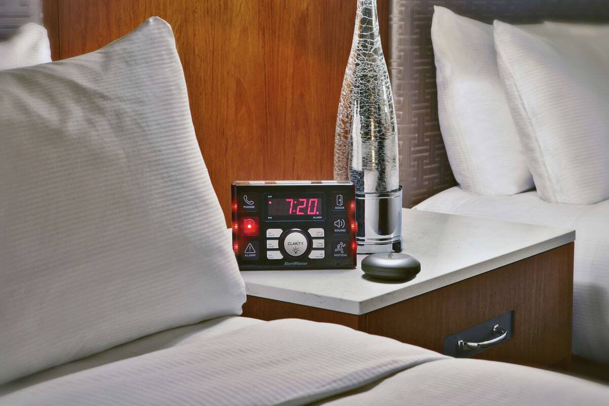 Una mesita de noche con un reloj digital que marca las 7:20, un control remoto y un objeto decorativo de vidrio.