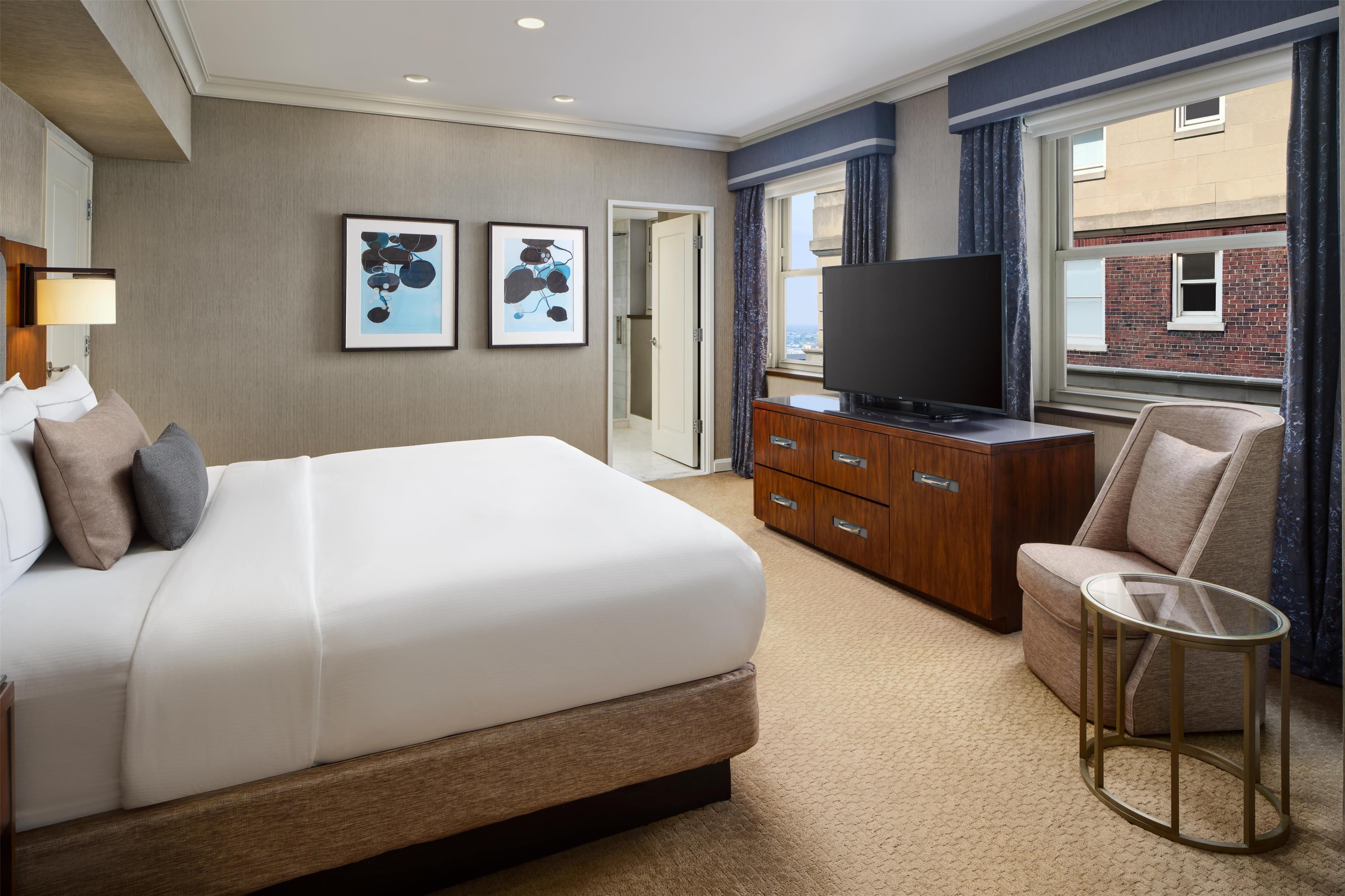 Ein Hotelzimmer mit einem großen Bett, einem Fernseher, einer Holzkommode und Blick auf ein Gebäude.