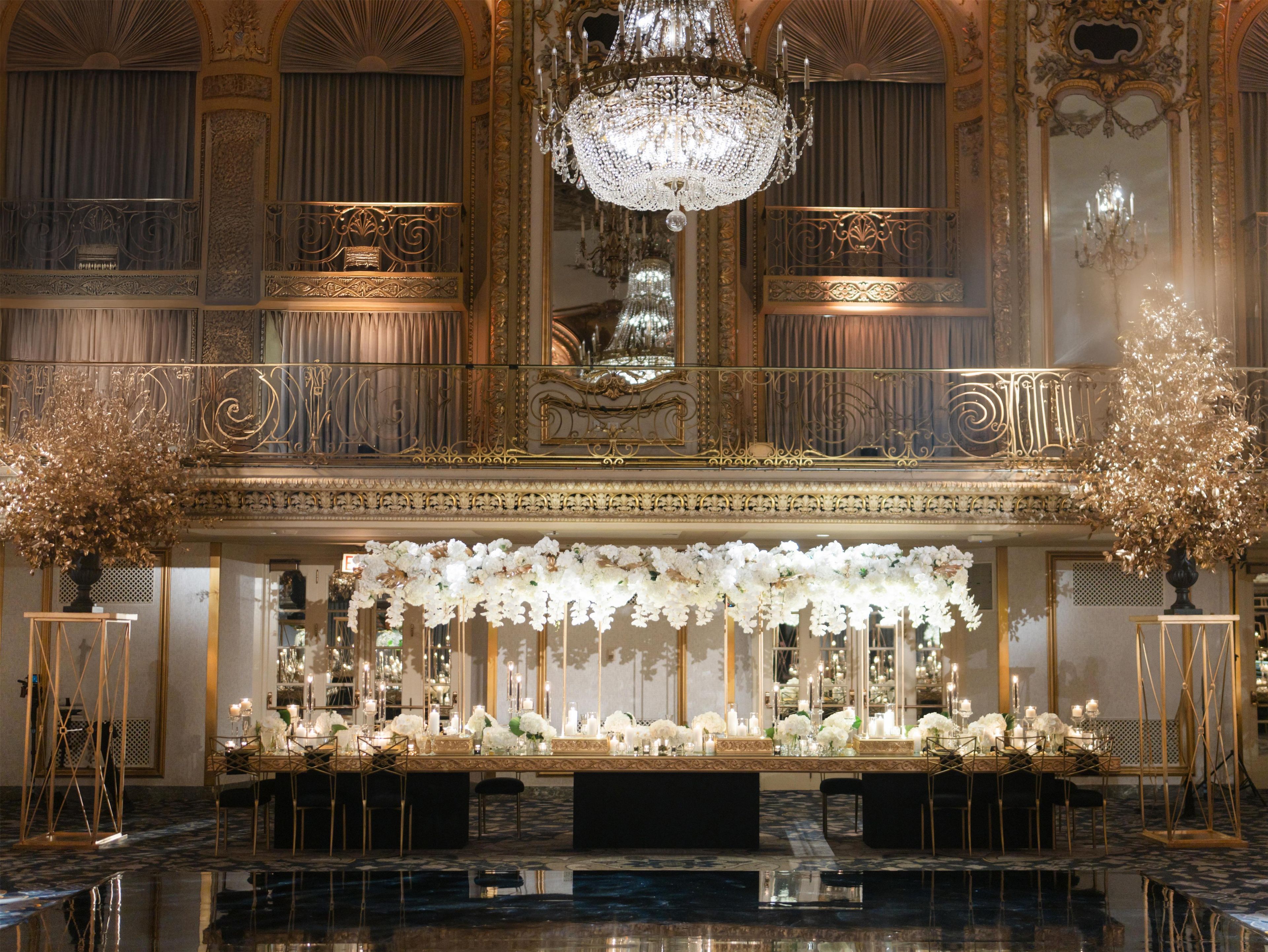 An opulent banquet hall with a grand chandelier, ornate gold detailing, and a long table adorned with white flowers and candles.