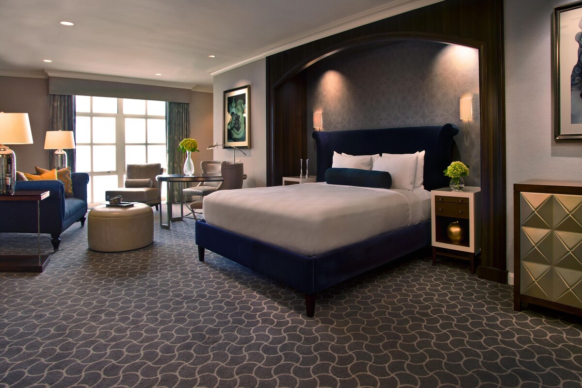 A hotel room with a large bed, a desk, a chair, a lamp, and a vase with flowers.