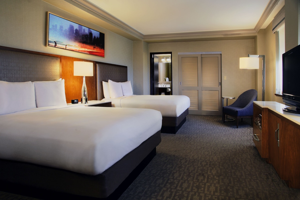 A hotel room with two beds, a desk with a television, a chair, a desk lamp, and a painting on the wall.