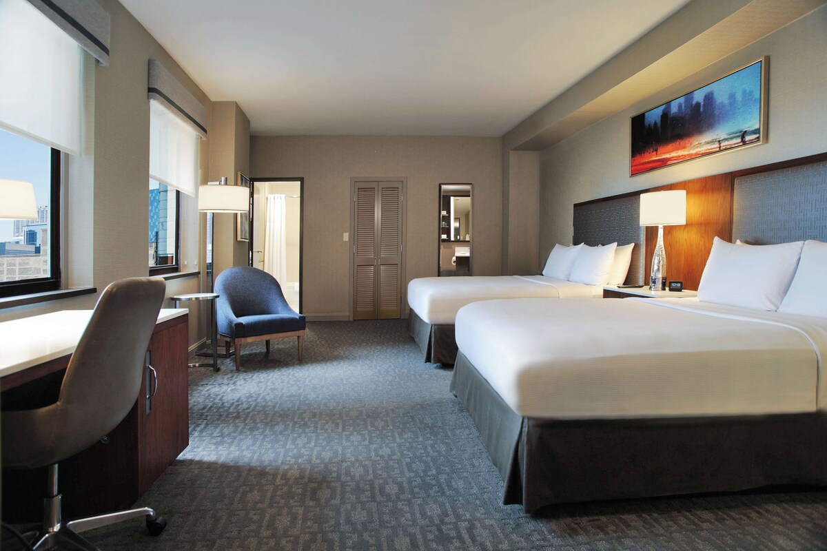 A hotel room with two beds, a desk, a chair, and a large window.