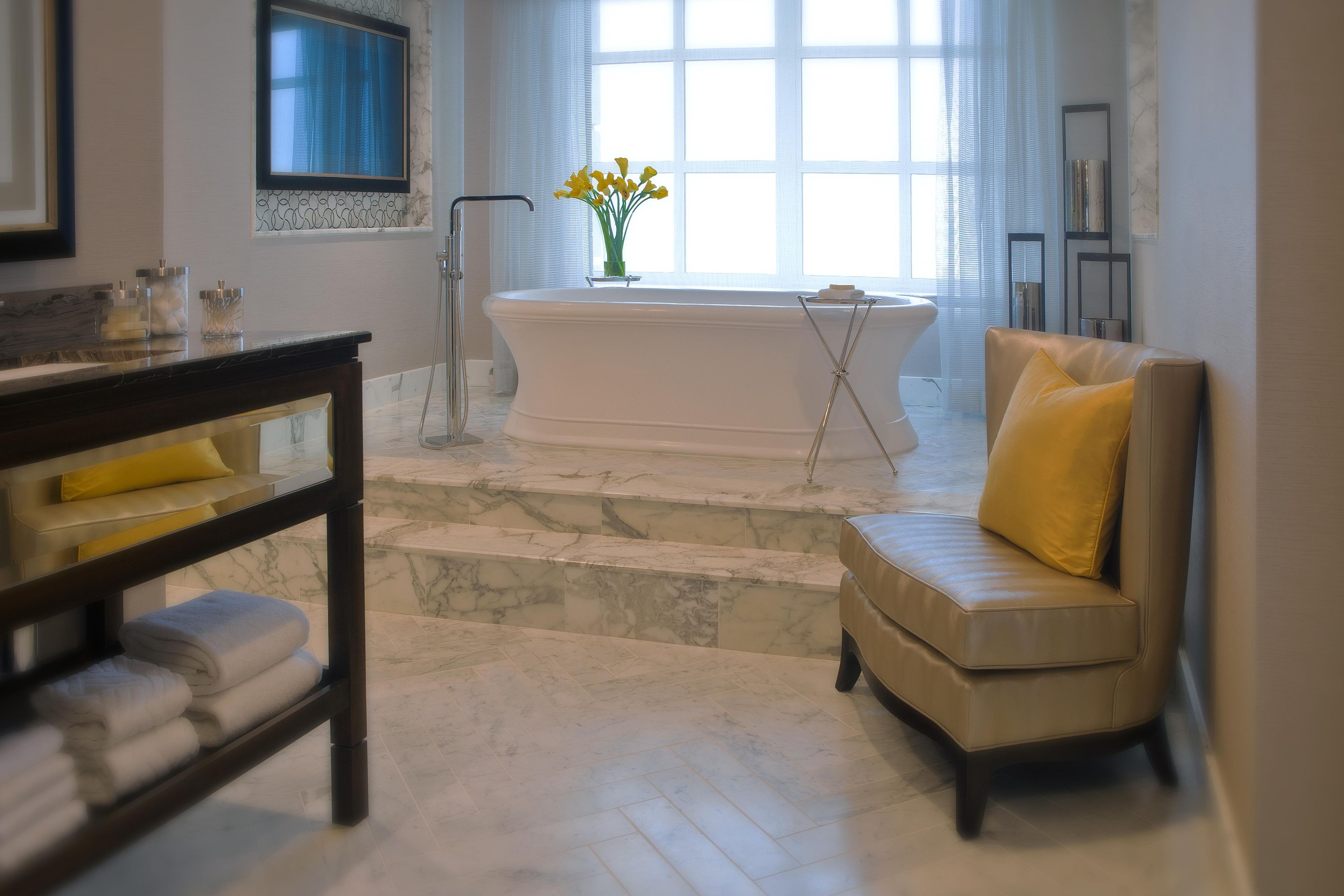 A bathroom with a bathtub, a vanity with a mirror, a chair, and a window with curtains.