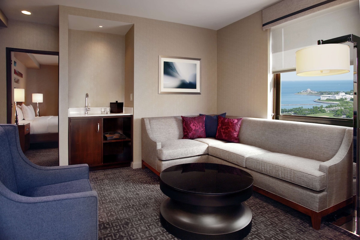 A hotel room with a sofa, a coffee table, a blue armchair, a desk with a lamp, a framed picture on the wall, and a window with a view of the sea.