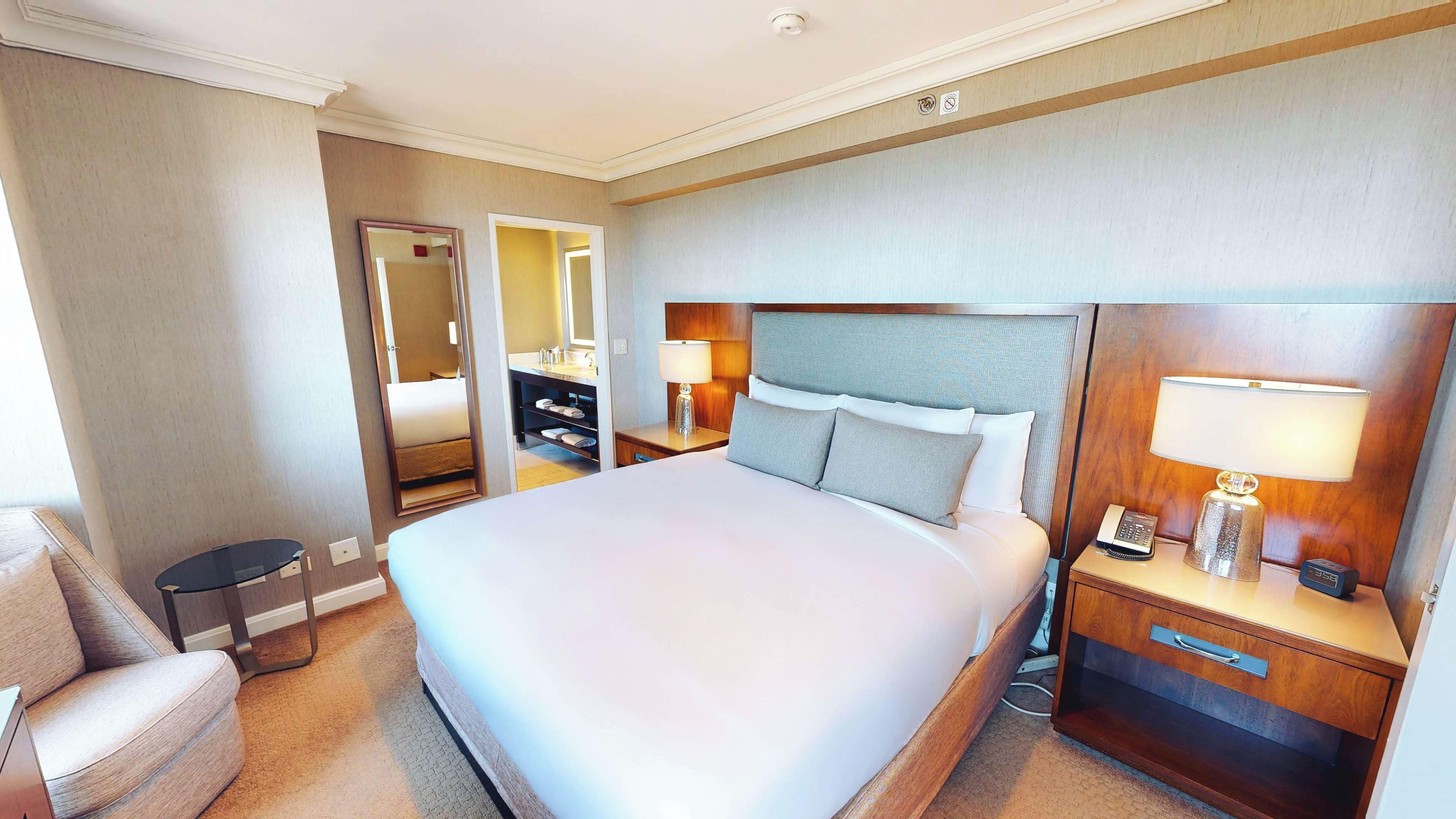 Una habitación de hotel con una cama grande, una mesita de noche con lámpara, un buró con teléfono, una silla, un aparador y un espejo.