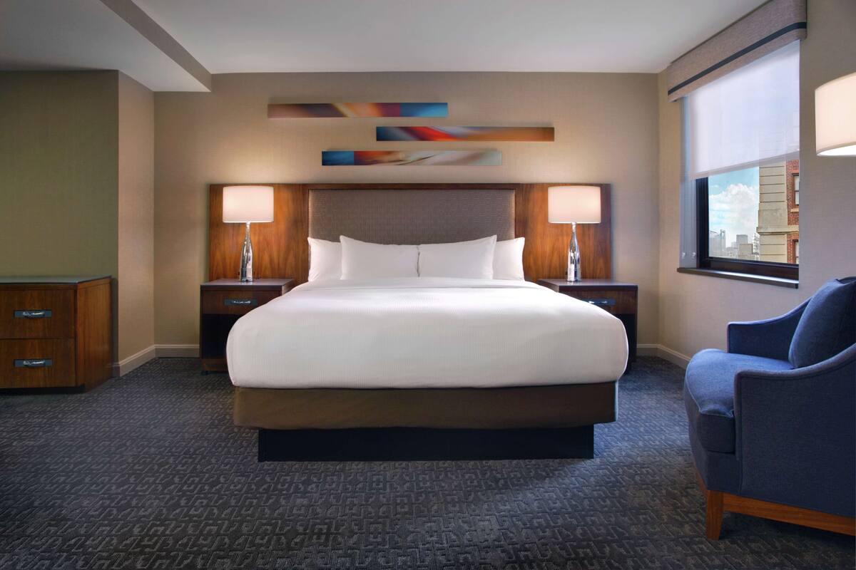 Una habitación de hotel con una cama grande, dos mesitas de noche con lámparas, un sillón azul y arte abstracto en la pared.