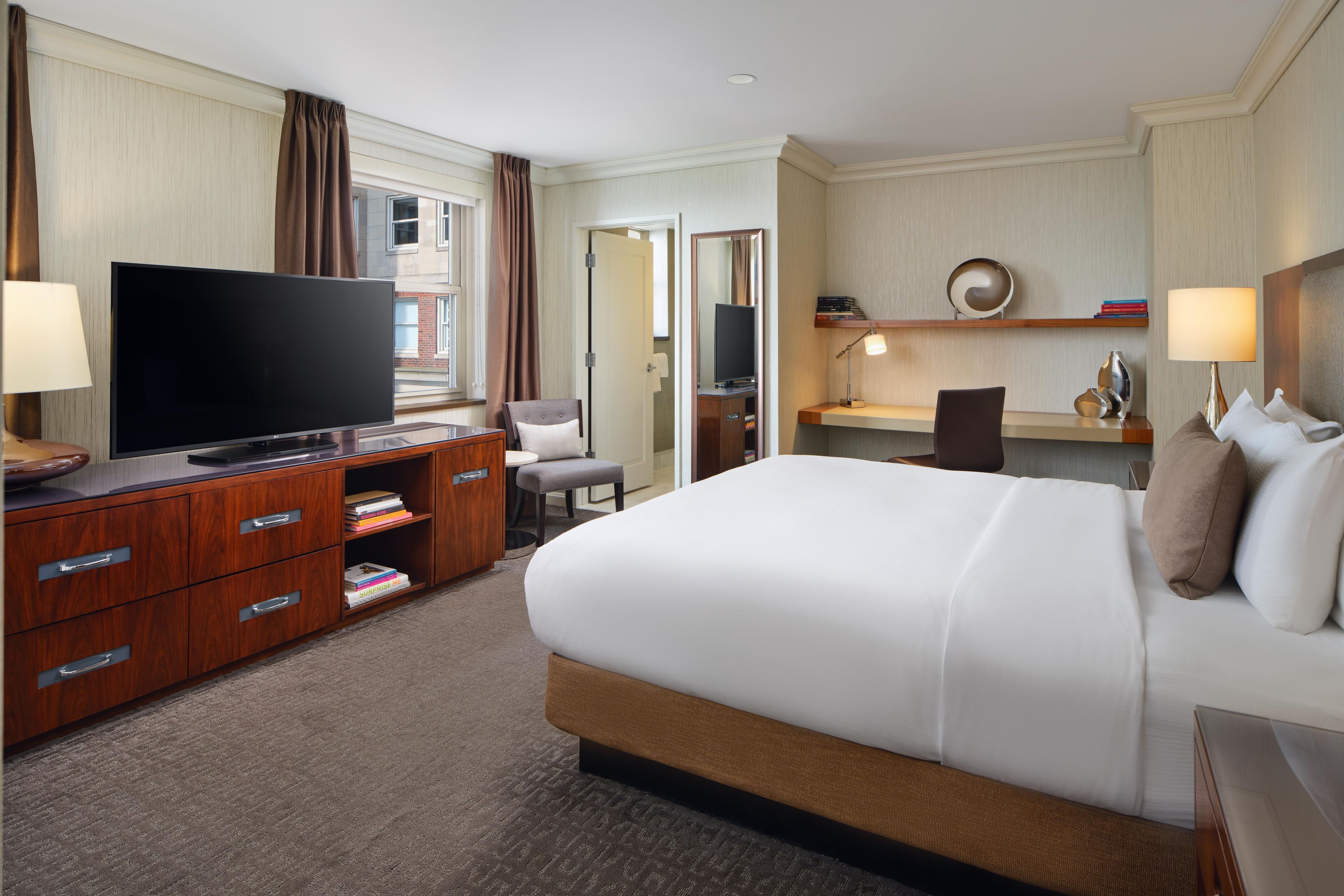 A hotel room with a large bed, a TV, a desk with a chair, and a bookshelf.