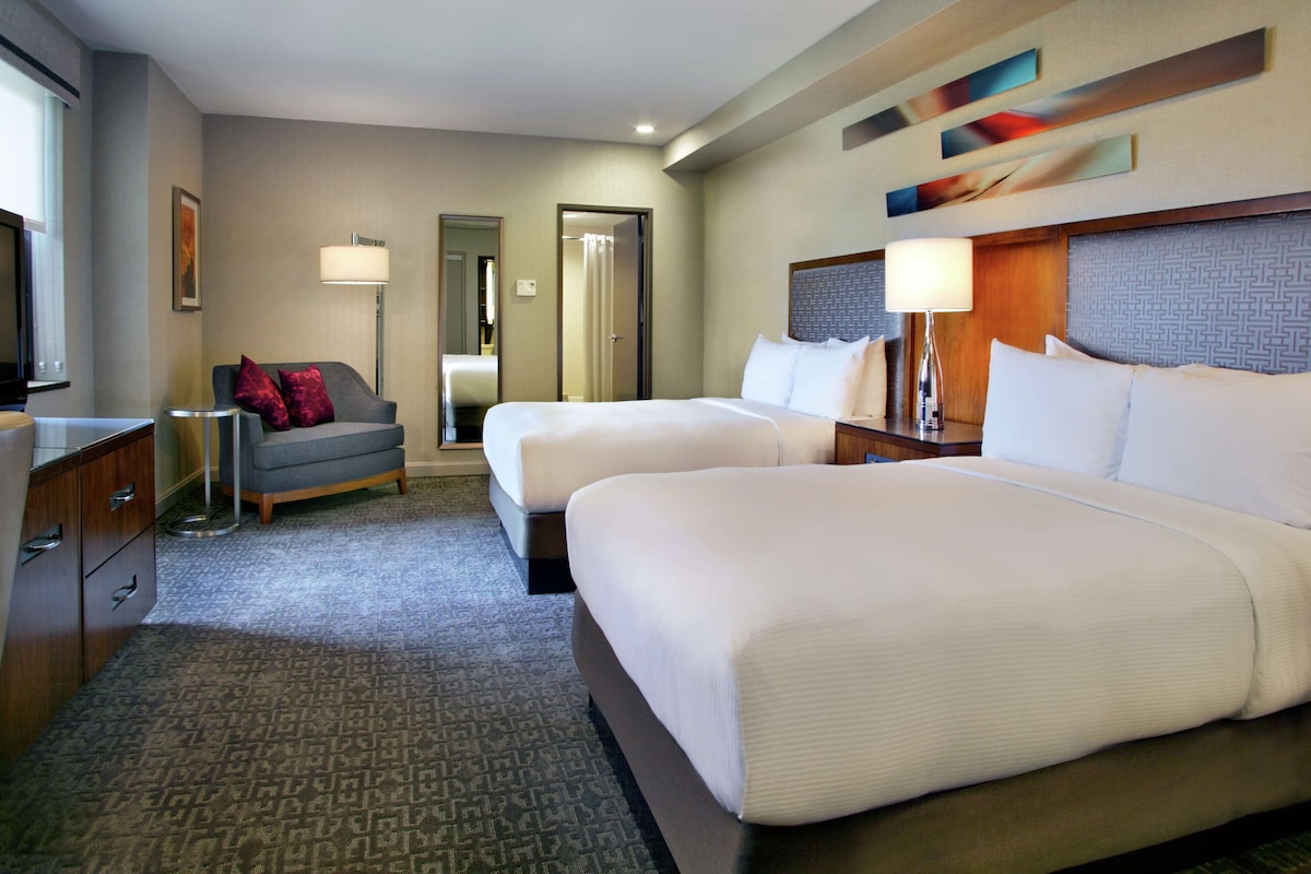 A hotel room with two beds, a desk, a chair, a lamp, and a mirror.