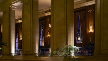 A grand lobby with large columns, ornate chandeliers, and a staircase leading to an upper level.