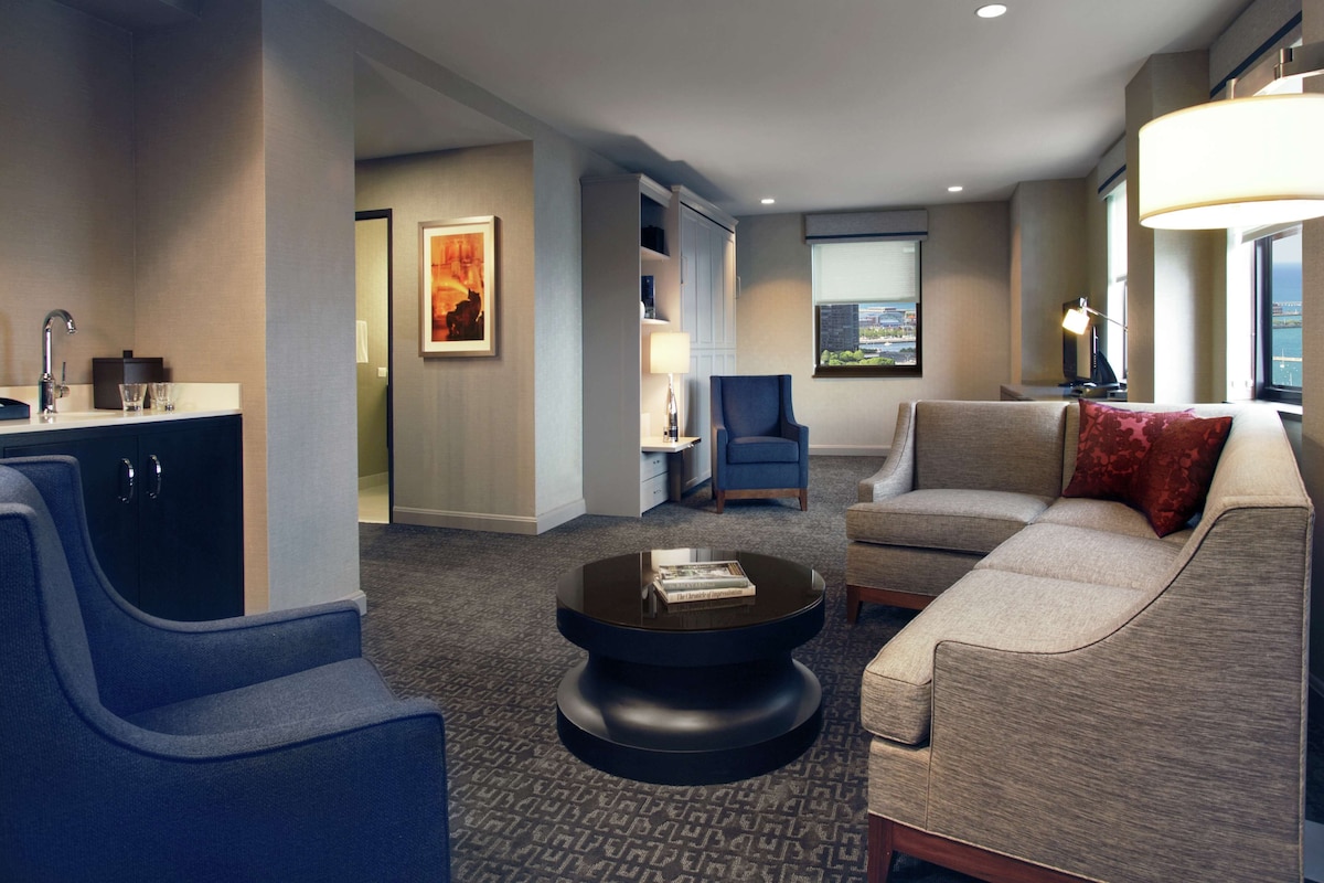 A modern hotel room with a sofa, armchair, and a round coffee table. There is a bookshelf and a desk with a lamp.