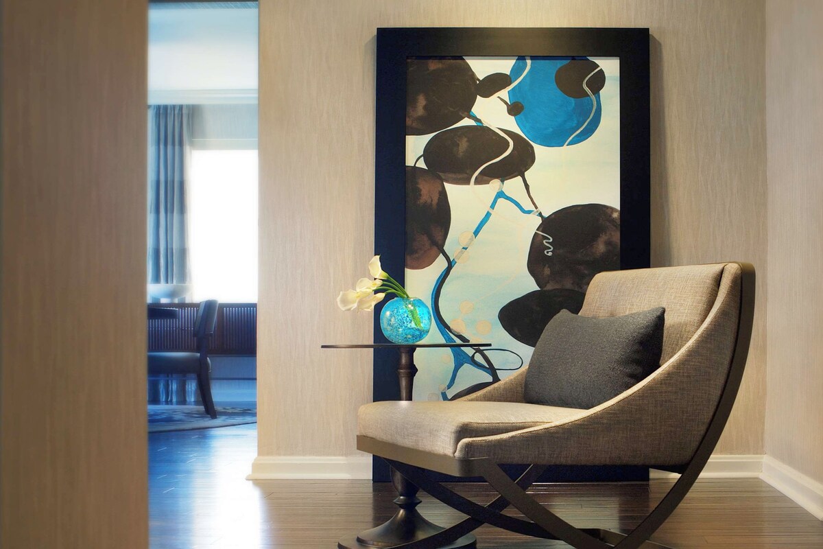 A modern interior with a curved upholstered chair, a glass table with a vase, and a colorful abstract painting on the wall.