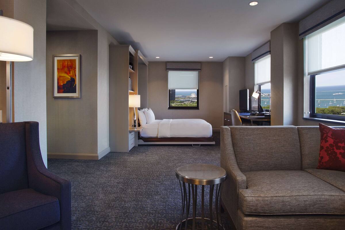 Habitación de hotel con cama, sofá, una mesita, escritorio, lámpara, un cuadro y ventana con vistas.