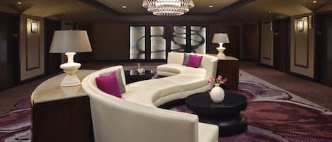 A modern hotel lobby with a curved white sofa, two side tables with lamps, a black coffee table, and a large chandelier.
