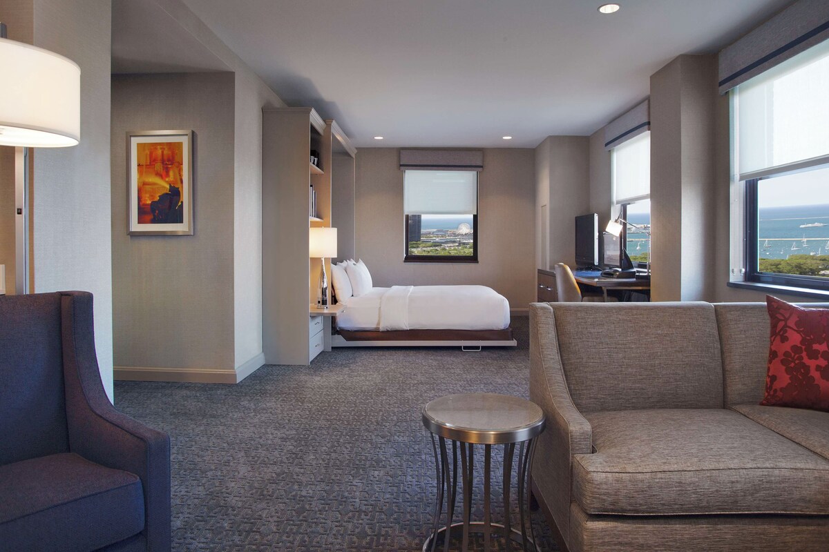 A hotel room with a bed, a sofa, a small table, a desk, a lamp, a painting, and a window with a view.