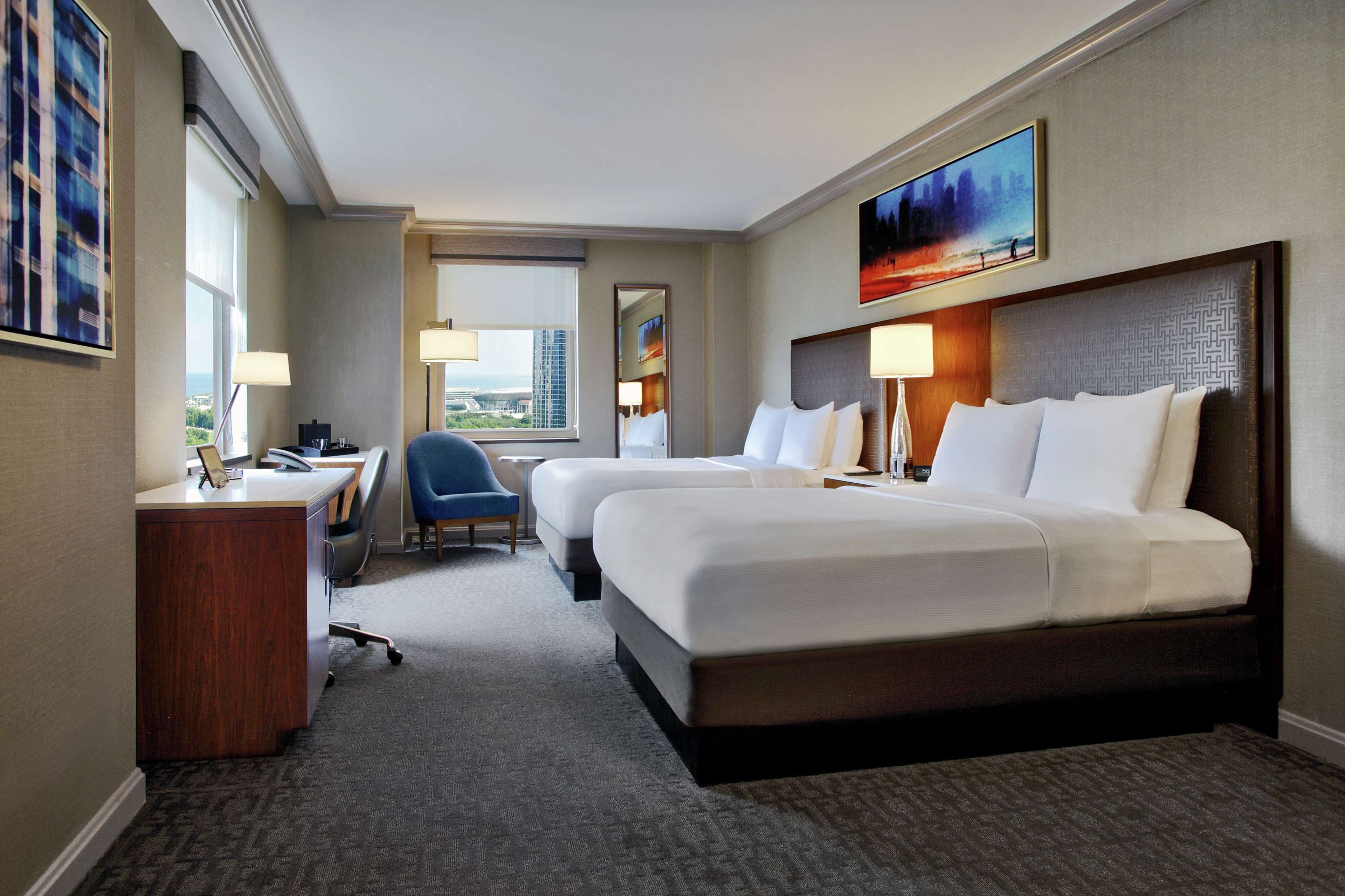 A hotel room with two beds, a desk, a chair, a window with a city view, and a painting on the wall.