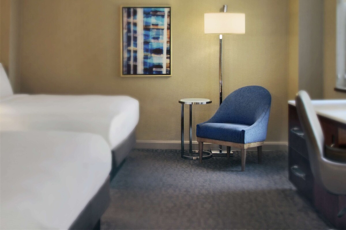 A hotel room with a bed, a blue chair, a small round table, and a floor lamp.