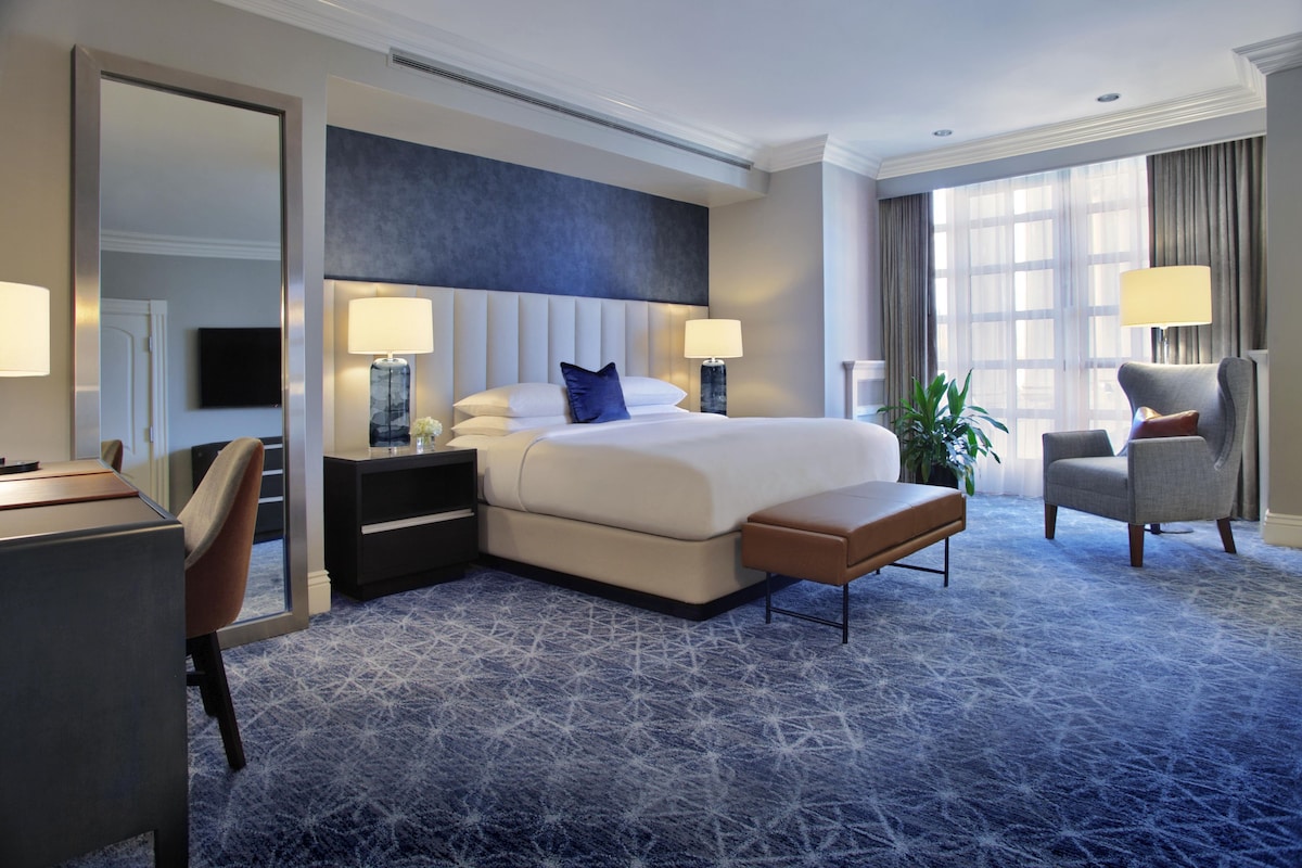 A hotel room with a large bed, bedside tables, a desk, a chair, and a large window.