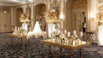 A grand ballroom with ornate chandeliers, a multi-tiered wedding cake centerpiece, and elegant floral arrangements on tables.