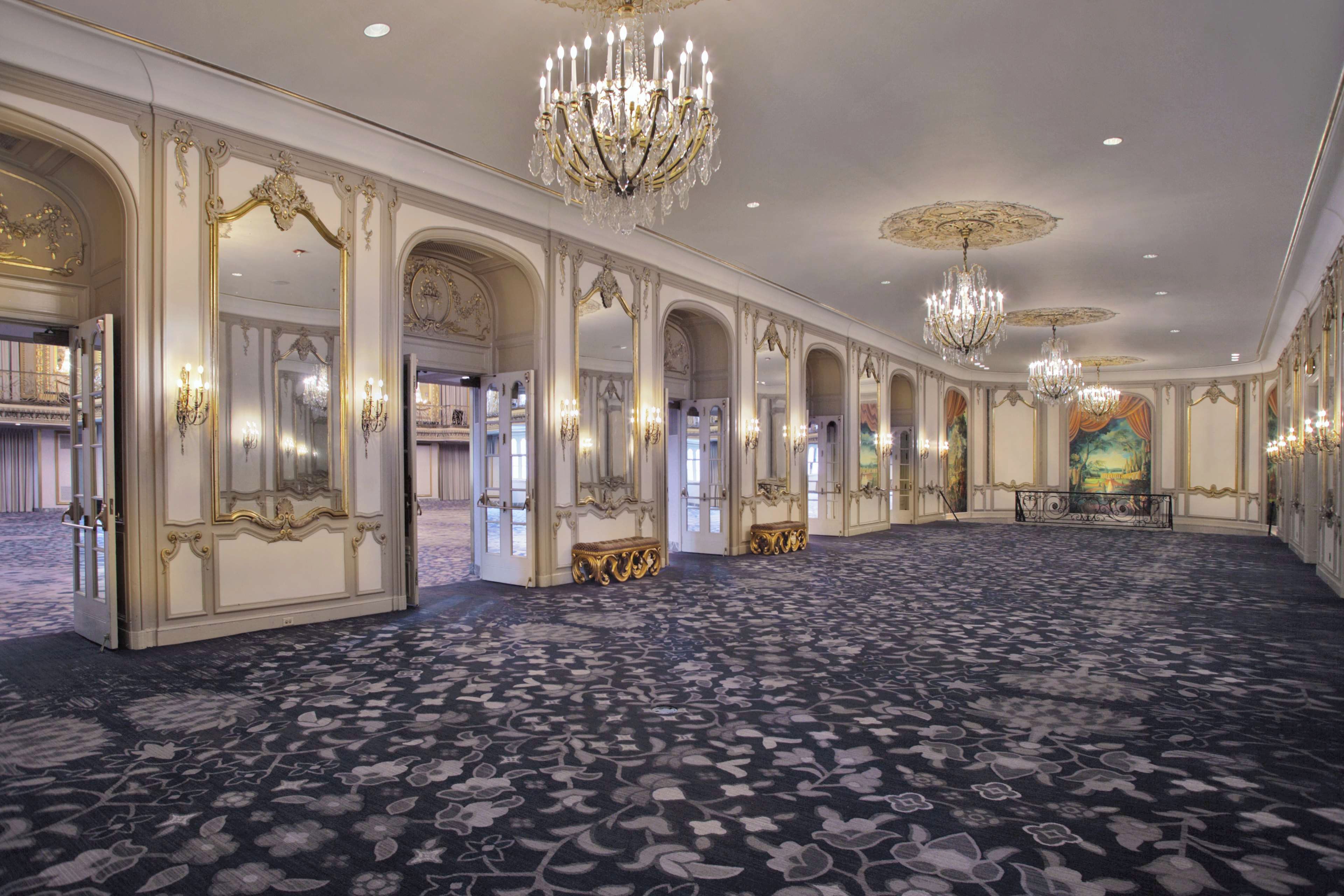 A grand hall with ornate chandeliers, high ceilings, and a patterned carpet.