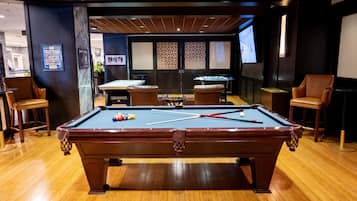 A pool table in a room with wooden flooring, two leather chairs, and a bar area with stools.