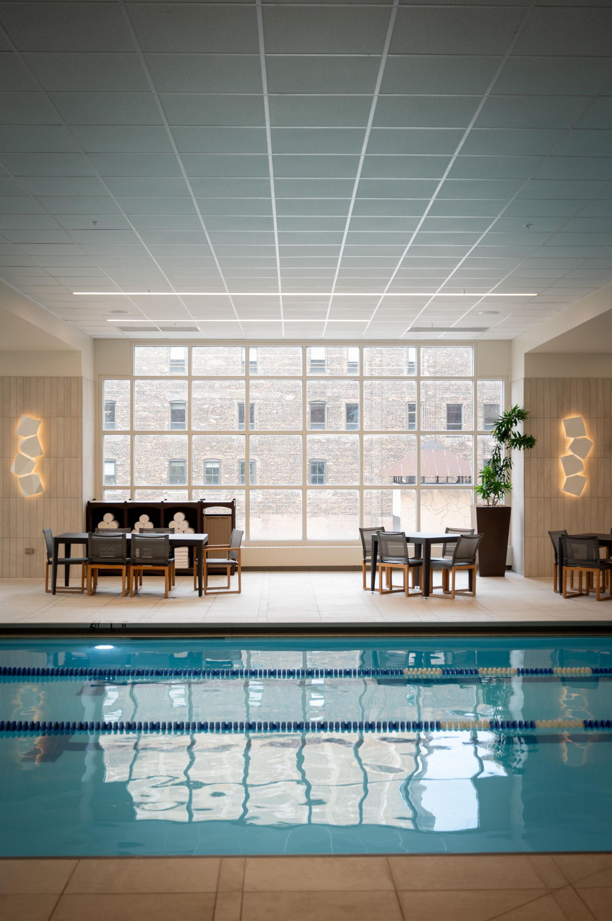 An indoor swimming pool with a view of a brick building through large windows. There are seating areas with tables and chairs.