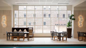 An indoor swimming pool with a view of a brick building through large windows. There are seating areas with tables and chairs.
