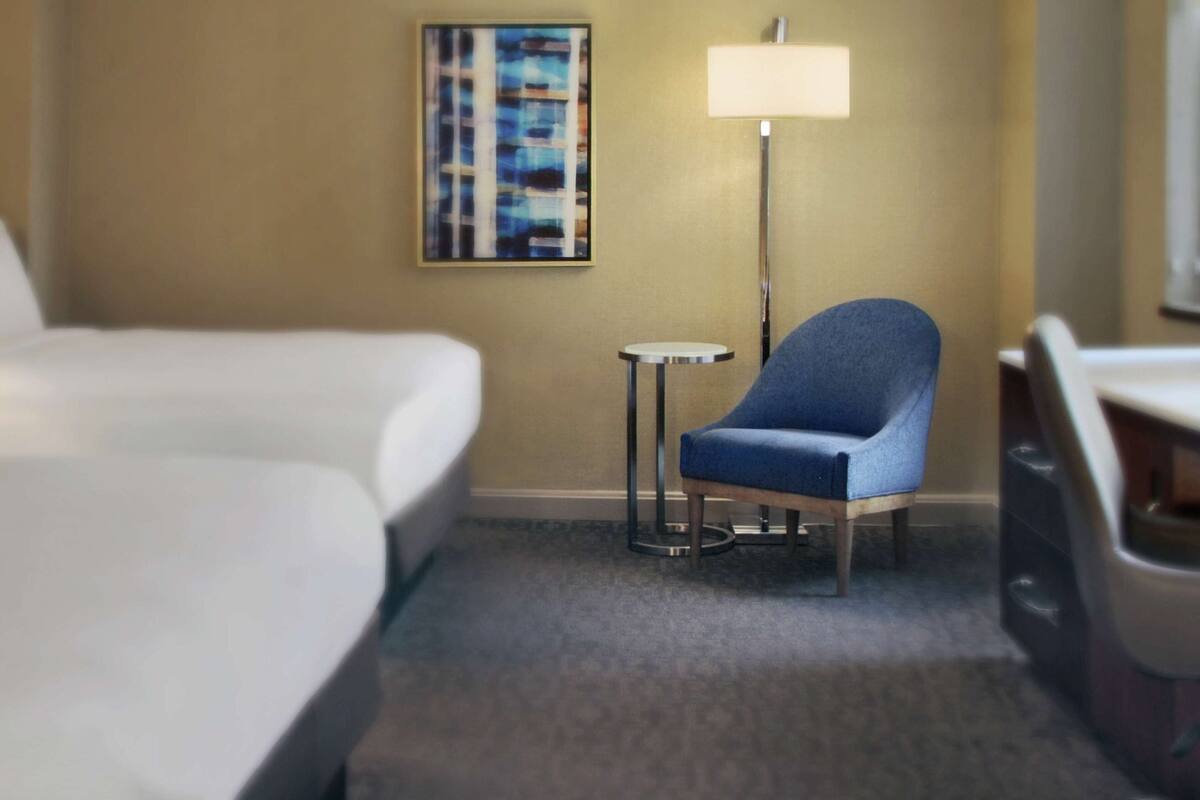 Una habitación de hotel con una cama, una silla azul, una mesita redonda y una lámpara de pie.