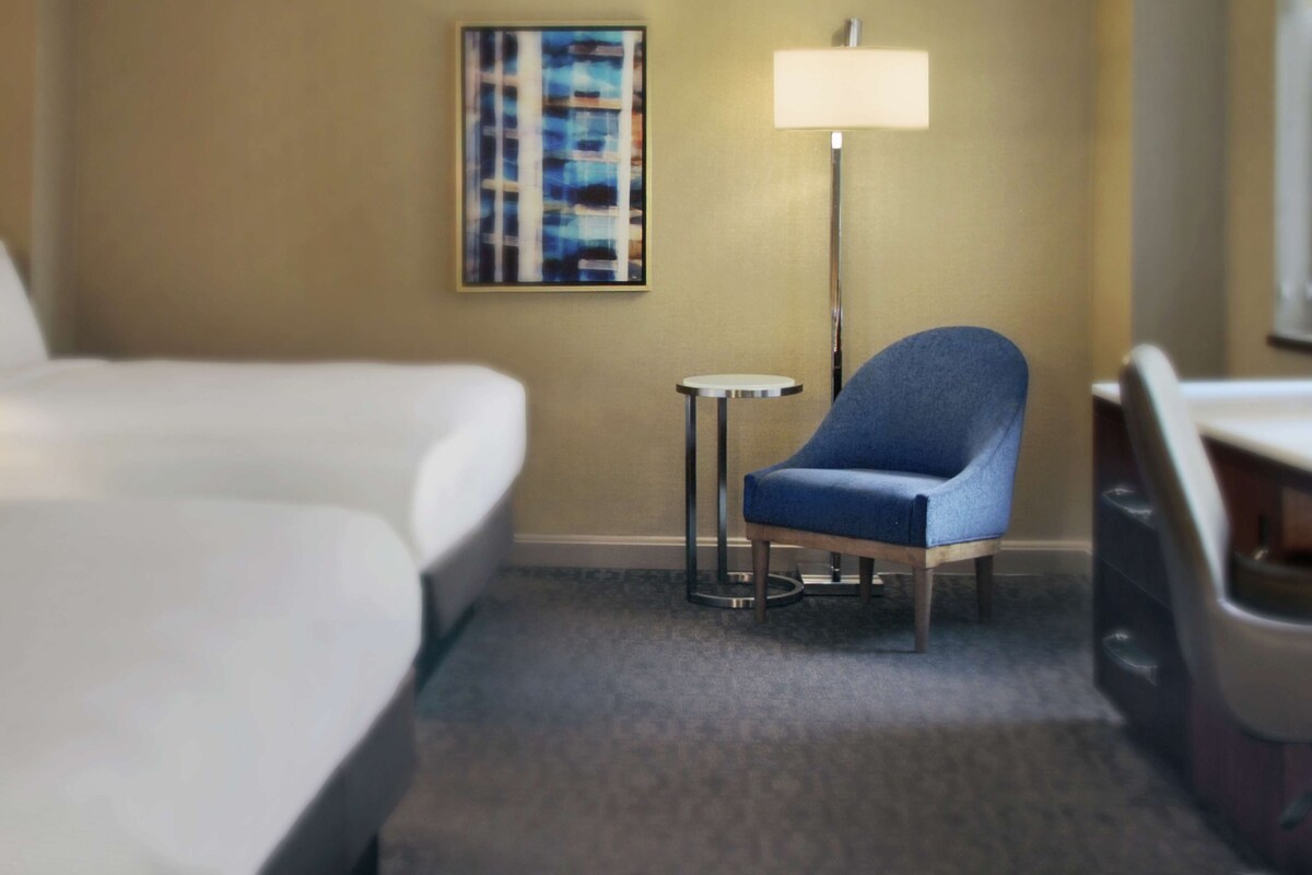Una habitación de hotel con una cama, una silla azul, una mesita redonda y una lámpara de pie.