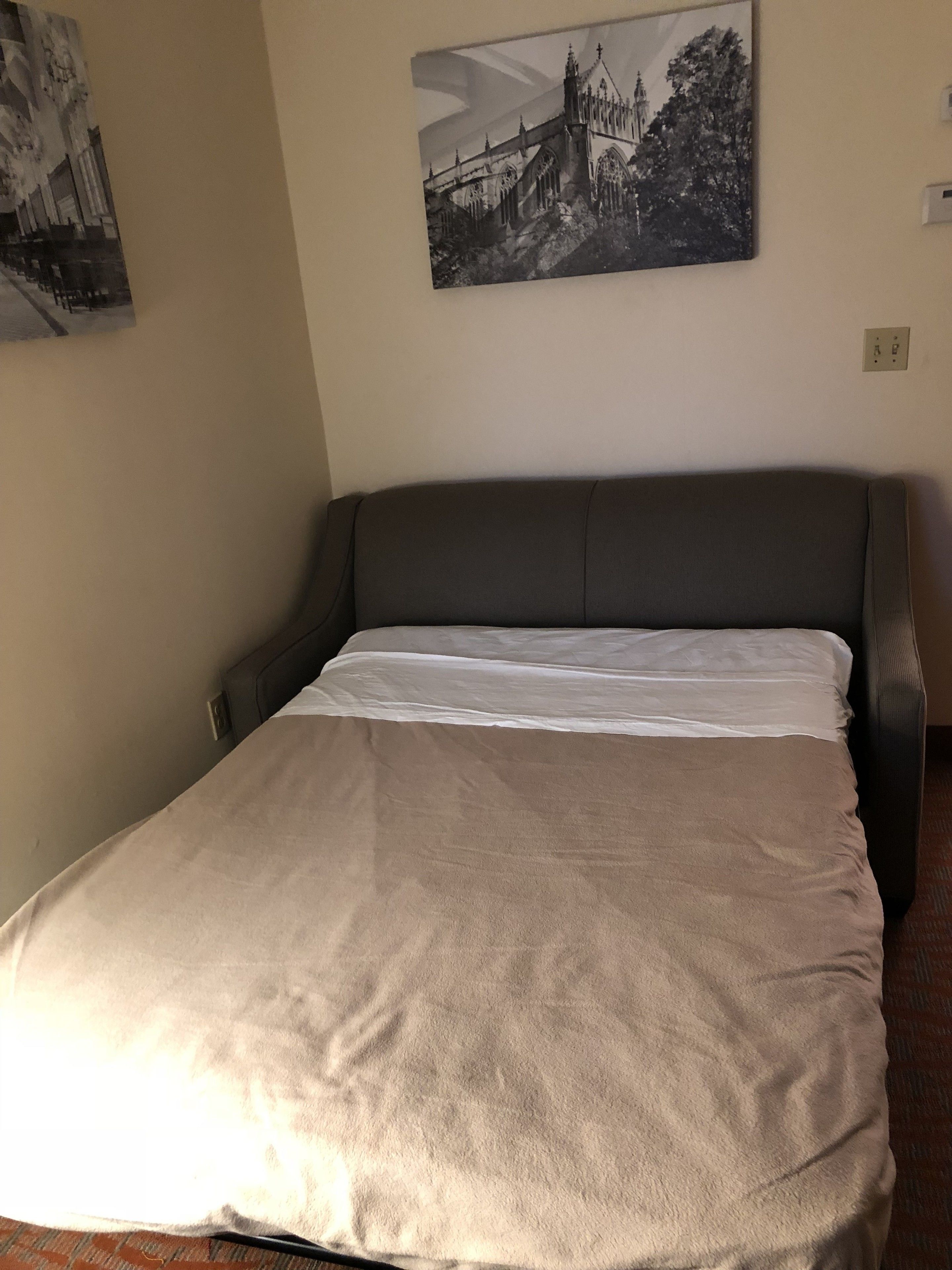 Una cama bien hecha con cabecera gris y manta color beige, un cuadro blanco y negro de un castillo en la pared, y un enchufe en la pared.