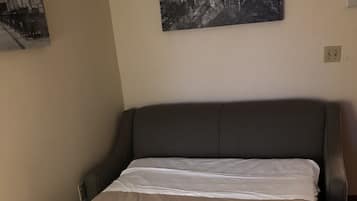Una cama bien hecha con cabecera gris y manta color beige, un cuadro blanco y negro de un castillo en la pared, y un enchufe en la pared.