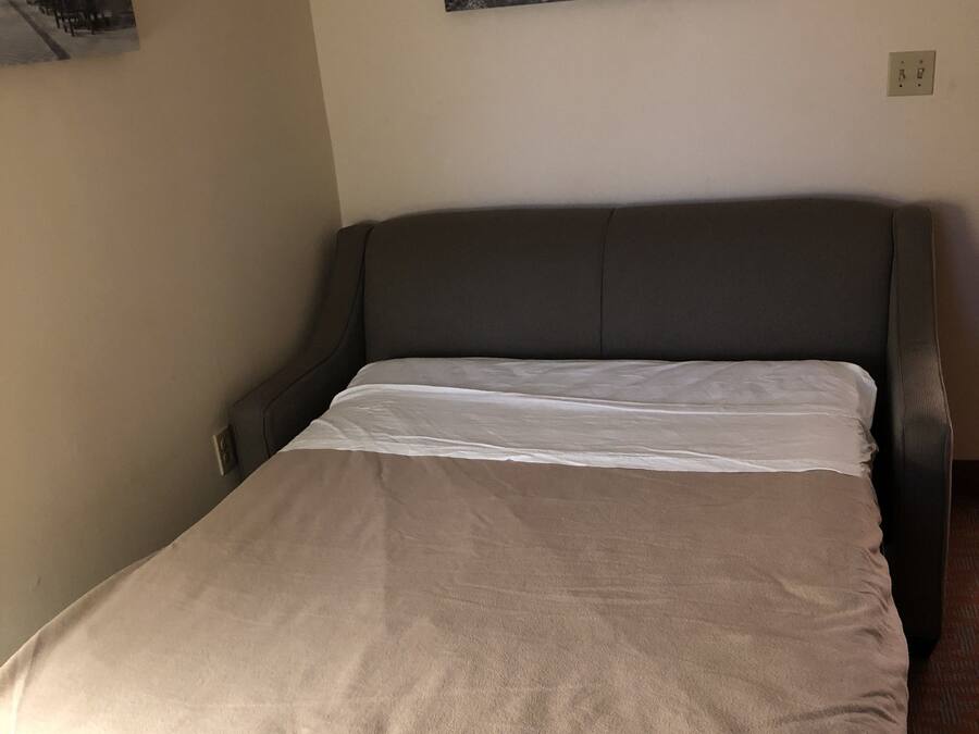 Una cama bien hecha con cabecera gris y manta color beige, un cuadro blanco y negro de un castillo en la pared, y un enchufe en la pared.