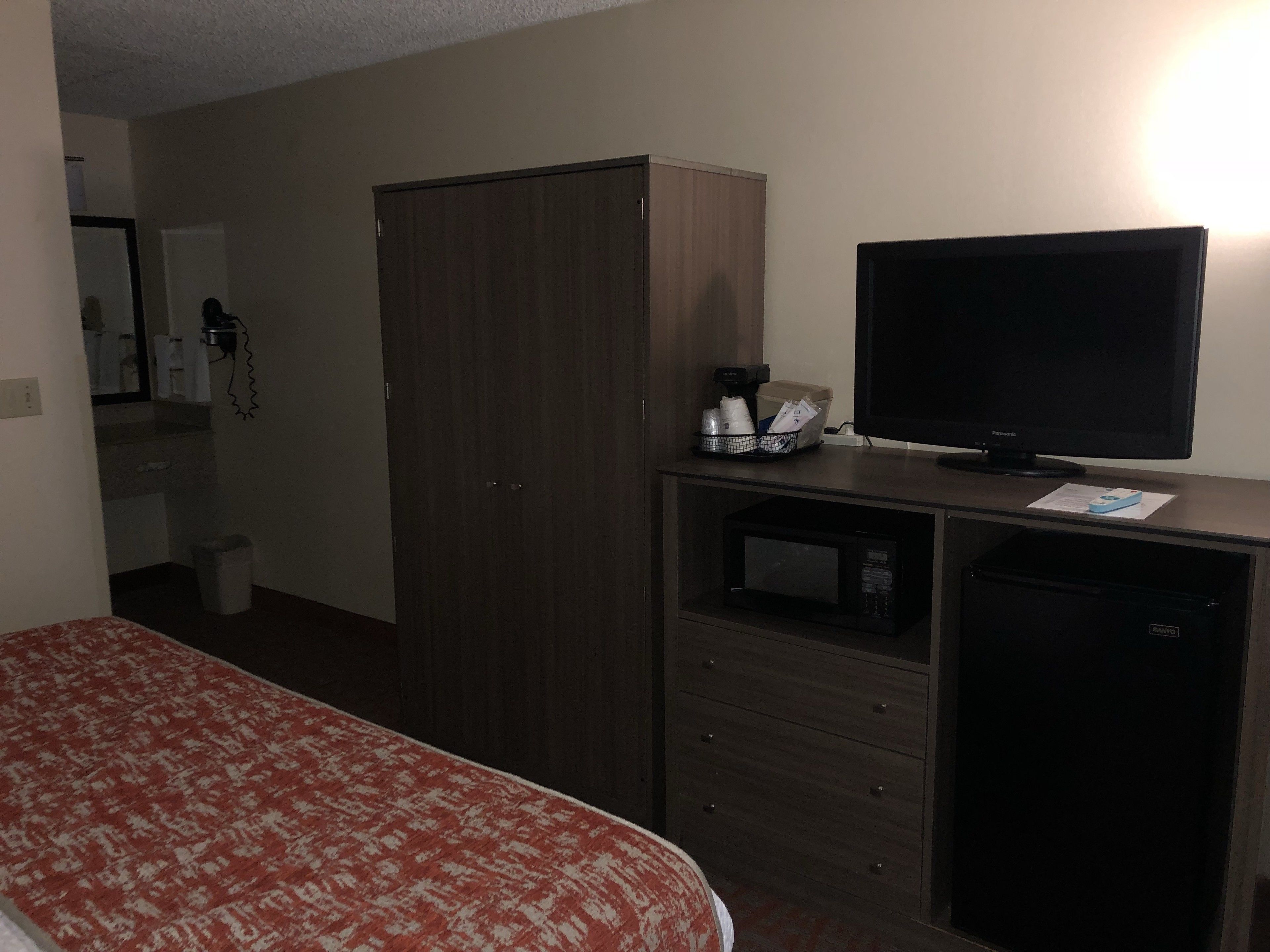 Una camera d'albergo con un letto, un armadio, una televisione, un forno a microonde e una scrivania con un computer e una tazza.