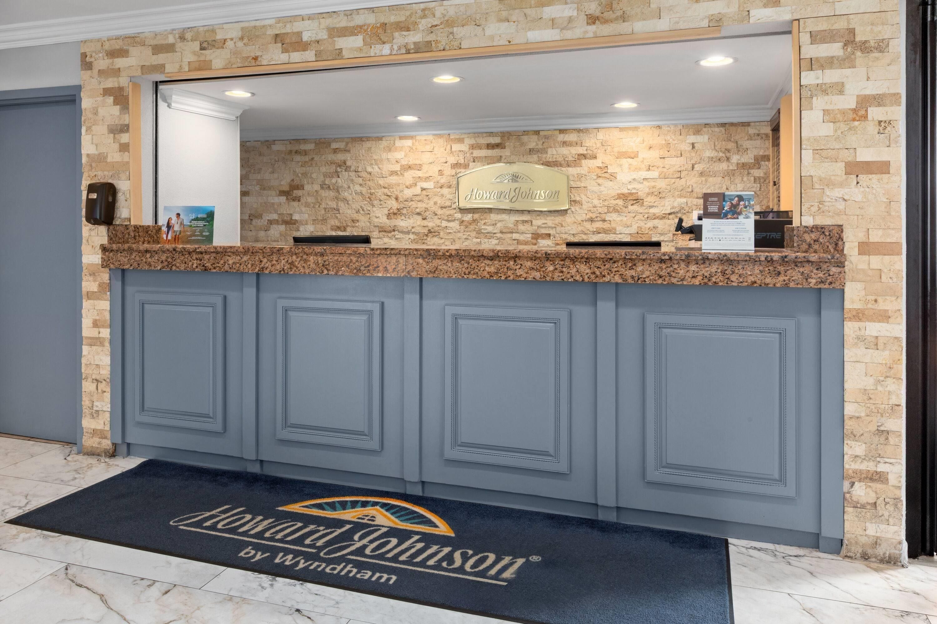 A reception desk with a stone wall backdrop, a nameplate, and a doormat with the hotel name.