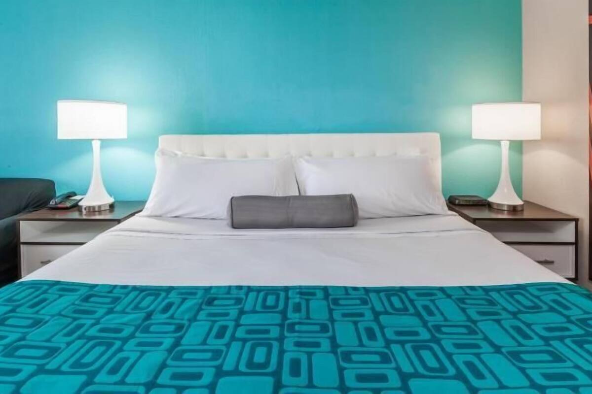 A hotel room with a bed, two bedside tables, a nightstand, a black sofa, and a teal-colored wall.