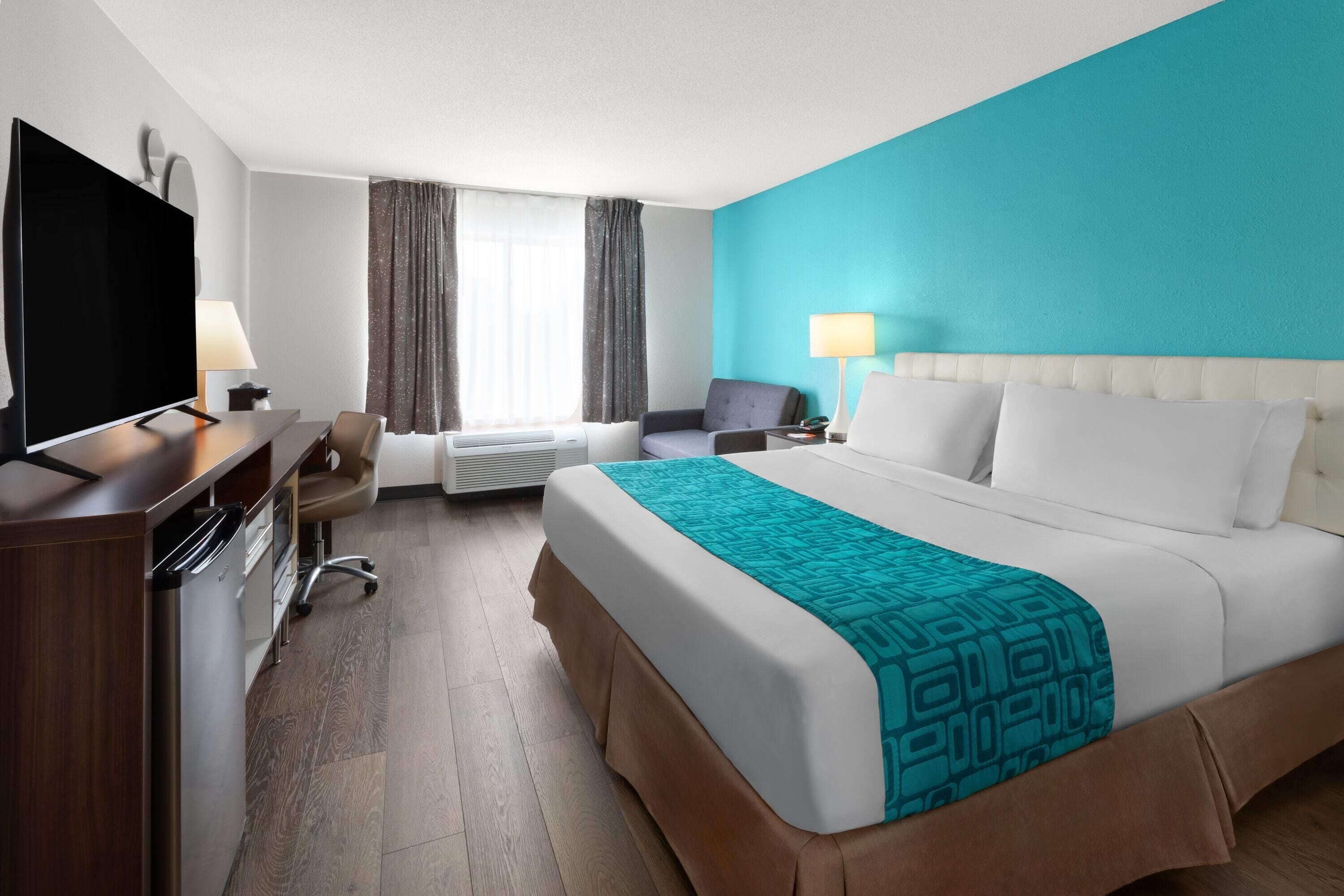 A hotel room with a bed, a desk with a TV, a chair, a lamp, and a window with curtains.