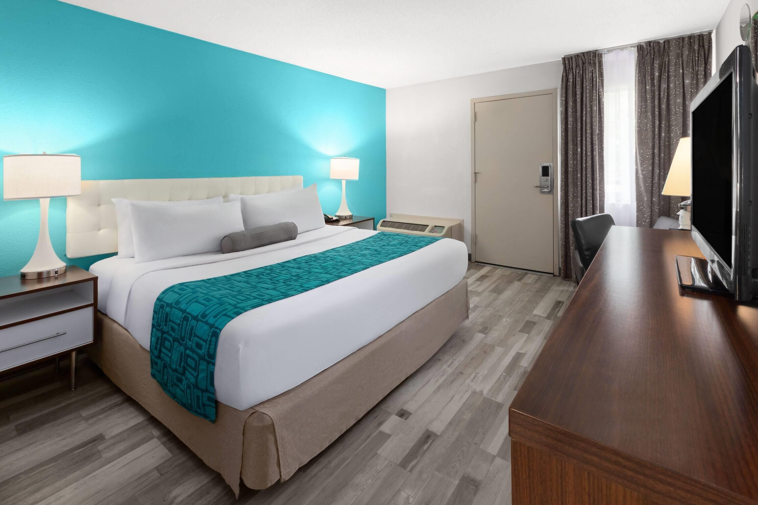 Quarto de hotel com uma cama grande, duas mesas de cabeceira com luminárias, uma escrivaninha com cadeira, uma televisão e uma porta com cortinas.