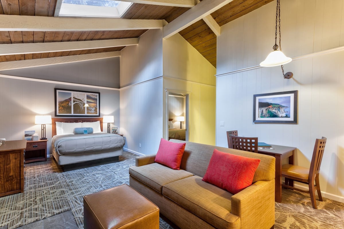 A modern hotel room with a bed, a sofa, a dining table, and chairs. There are two lamps on the nightstands and a large window with a view.