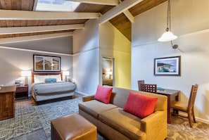 A modern hotel room with a bed, a sofa, a dining table, and chairs. There are two lamps on the nightstands and a large window with a view.