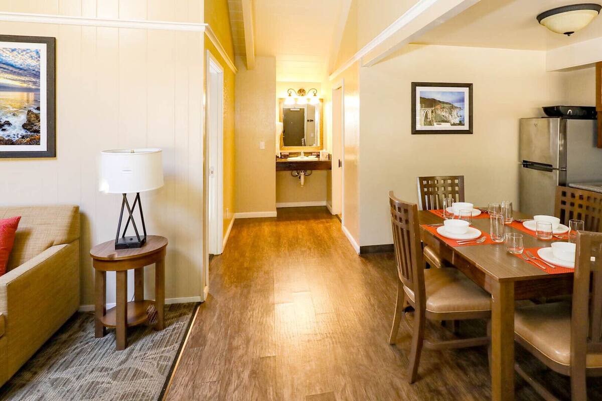 A dining area with a wooden table set for four, a kitchen with wooden cabinets, and a hallway leading to a bathroom.
