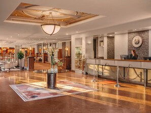 Un vestíbulo de hotel con un mostrador de recepción, una mesa de centro de vidrio con flores y una araña.