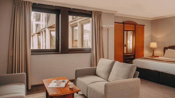 Habitación de hotel con sofá, una mesita con una revista y una taza, cama con mesita de noche, lámpara y vista de edificios a través de la ventana.