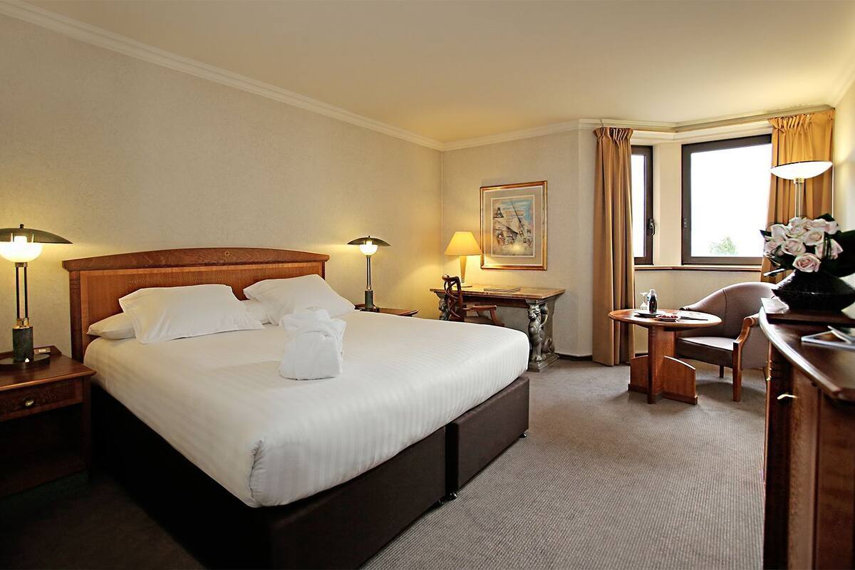 A hotel room with a large bed, bedside lamps, a desk, a chair, and a window with curtains.