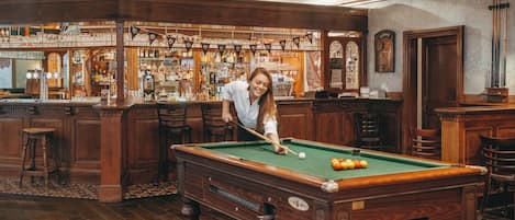 Une table de billard dans un bar, avec des meubles en bois et un comptoir bien garni de bouteilles.