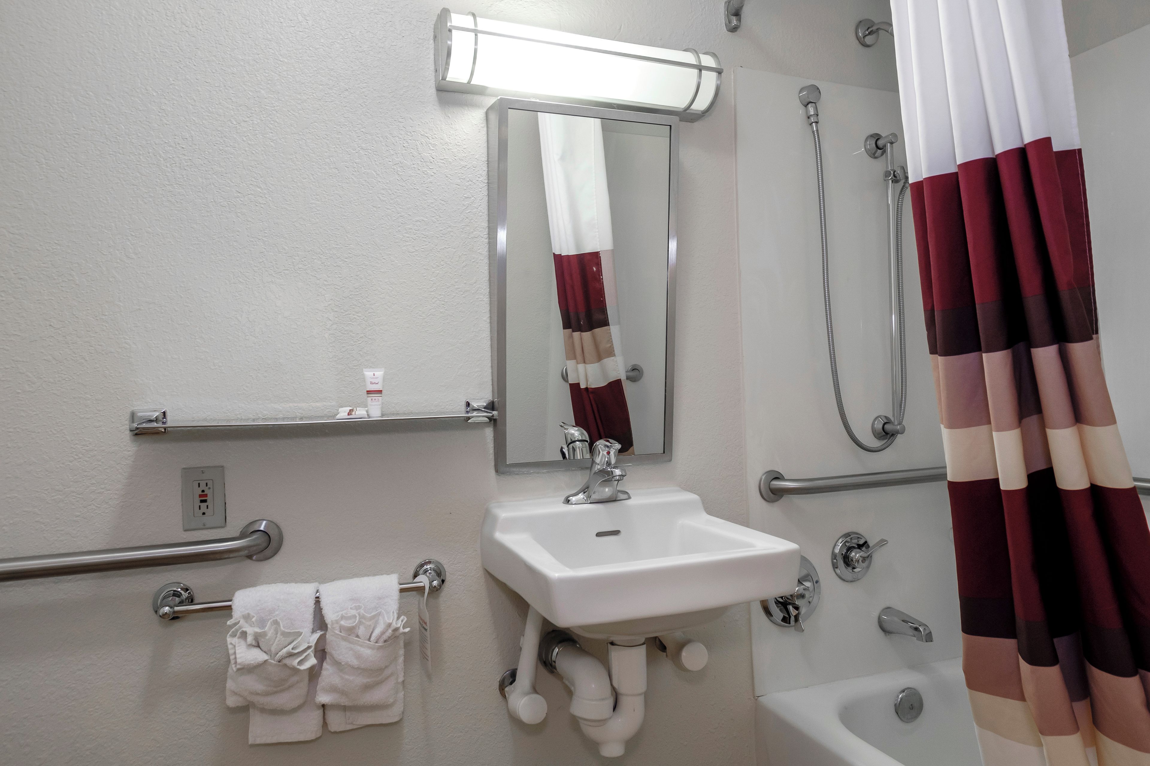 A bathroom with a sink, mirror, shower, and handrails.