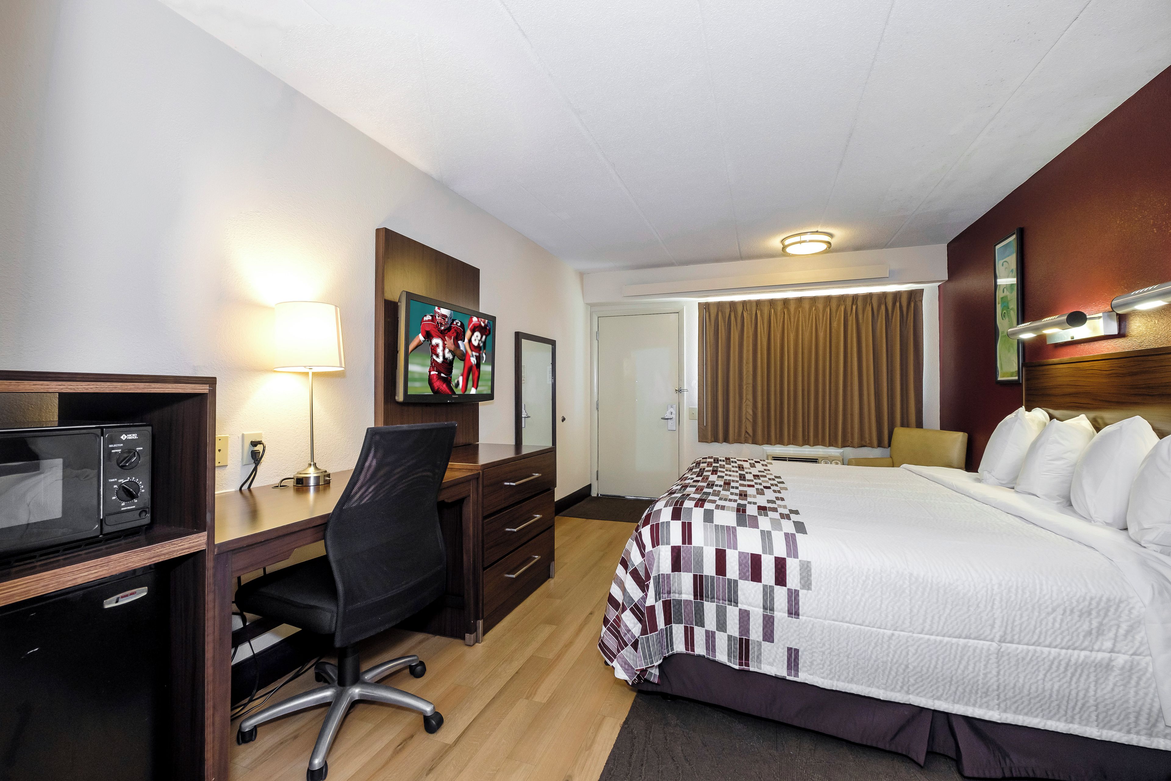 A hotel room with a bed, desk, chair, microwave, and artwork on the wall.
